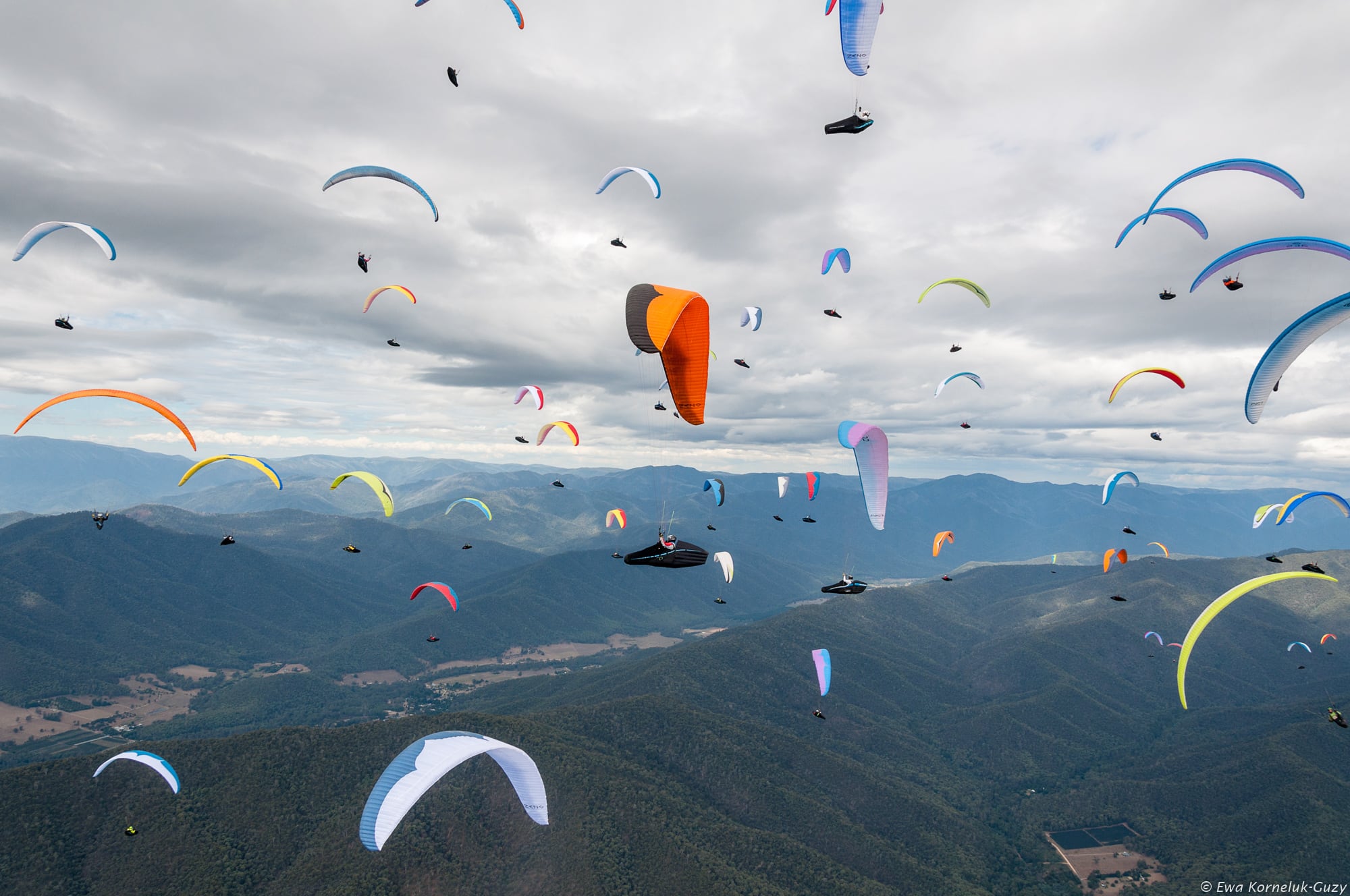 تعداد زیادی پاراگلایدر رنگارنگ در آسمان ابری بر فراز کوه‌ها و دره‌های سرسبز در حال پرواز هستند، منظره‌ای پر از انرژی و هیجان در یک رقابت یا تجمع بزرگ پرواز با پاراگلایدر